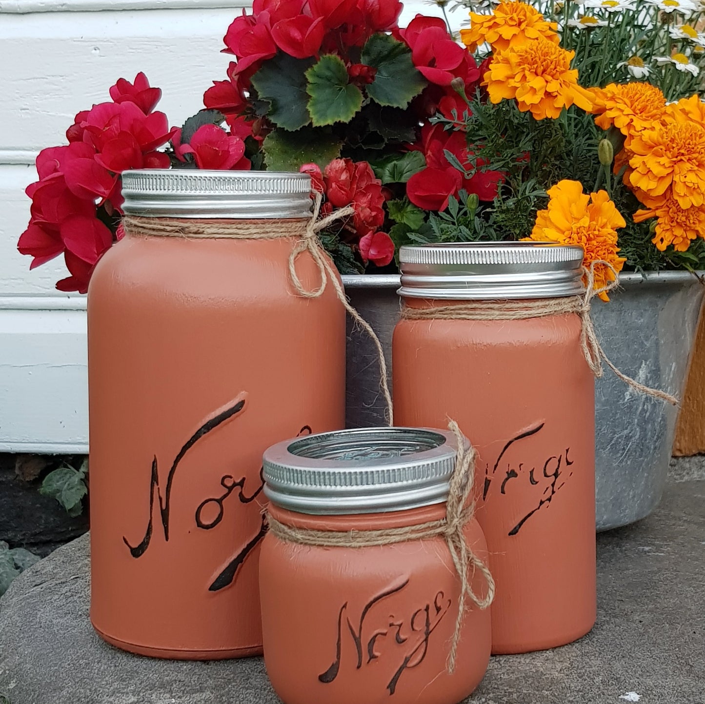 Tre norgesglass i ulik størrelse malt med fargen rusty, blomster i rødt og orange i en metallboks bak glassene